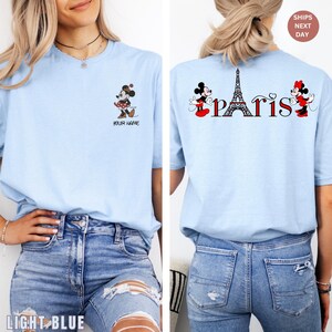 May include: Light blue t-shirt featuring Minnie Mouse and the word "Paris" with the Eiffel Tower. The front of the shirt has a Minnie Mouse graphic and the text "Your Name". The back of the shirt has the word "Paris" with Minnie Mouse graphics.