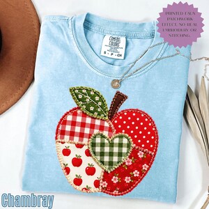 May include: A light blue t-shirt featuring a large apple graphic composed of various red, white, and green patterned sections including gingham, polka dots, and floral prints. A central green and white gingham heart sits on the apple.