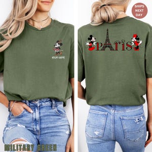 May include: Military green t-shirt featuring a cartoon Minnie Mouse design on the front and back. The back of the shirt has the word "Paris" with the Eiffel Tower and Mickey and Minnie Mouse. The front of the shirt has the text "YOUR NAME".