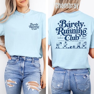 May include: A light blue t-shirt featuring a small dark blue logo on the front chest that reads Barely Running Club. The back of the shirt displays a large dark blue graphic with the text Barely Running Club and cartoon figures running.