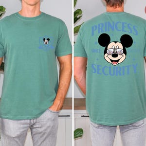 May include: A teal green t-shirt featuring a Mickey Mouse graphic. The front pocket has a small Mickey Mouse design. The back of the shirt displays "PRINCESS SECURITY" with Mickey wearing sunglasses. The shirt is a comfortable, casual style.