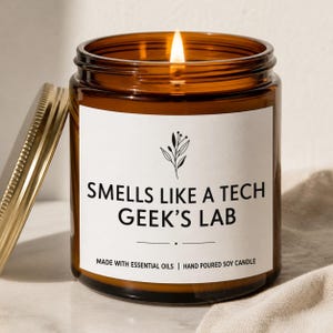 May include: A lit candle in an amber glass jar with a gold lid. The white label features a black botanical illustration and the text: SMELLS LIKE A TECH GEEK'S LAB, MADE WITH ESSENTIAL OILS | HAND POURED SOY CANDLE. The candle has a single flame.