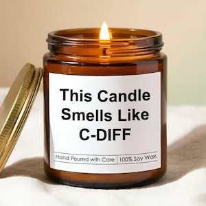 May include: A lit brown glass jar candle with a gold lid. The candle has a white label with the text "This Candle Smells Like C-DIFF". The label also reads "Hand Poured with Care 100% Soy Wax".
