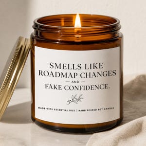 May include: An amber glass jar candle with a lit wick and a gold-colored metal lid. The white label features black text that reads: SMELLS LIKE ROADMAP CHANGES AND FAKE CONFIDENCE. MADE WITH ESSENTIAL OILS | HAND POURED SOY CANDLE.
