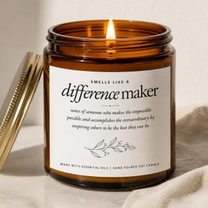 May include: A lit soy candle in a brown glass jar with a gold lid. The label reads "Smells Like a Difference Maker" with a description and botanical illustration. The candle is hand-poured with essential oils.