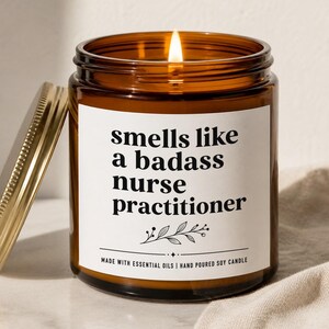 May include: A lit soy candle in an amber glass jar with a gold lid. The white label features black text that reads: smells like a badass nurse practitioner, with a small botanical illustration and the text: MADE WITH ESSENTIAL OILS | HAND POURED SOY CANDLE.