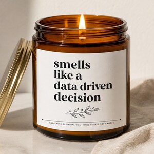 May include: A lit soy candle in a brown glass jar with a gold lid. The white label reads "smells like a data driven decision." The candle is hand-poured and made with essential oils.