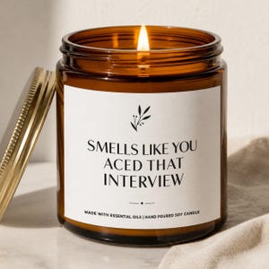 May include: A lit soy candle in an amber glass jar with a gold lid. The white label features a black botanical icon and the text: SMELLS LIKE YOU ACED THAT INTERVIEW, followed by MADE WITH ESSENTIAL OILS | HAND POURED SOY CANDLE.