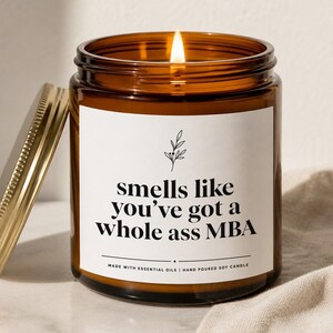 May include: A lit soy candle in an amber glass jar with a gold lid. The white label features a black botanical illustration and the text: smells like you've got a whole ass MBA. Below the text, it reads: MADE WITH ESSENTIAL OILS | HAND POURED SOY CANDLE.