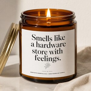 May include: A lit soy candle in an amber glass jar with a gold lid. The white label features black text reading: Smells like a hardware store with feelings. Below the text is a small botanical line drawing and the words Made with essential oils.