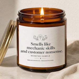 May include: A lit scented candle in an amber glass jar with a gold lid. The white label features a floral icon and the text: Smells like mechanic skills and customer nonsense. SCENTED CANDLE. Made with essential oils | hand poured soy candle.