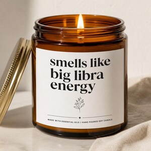 May include: A lit candle in an amber glass jar with a gold metal lid. The white label features black text that reads: smells like big libra energy, with a small floral illustration and the text: MADE WITH ESSENTIAL OILS | HAND POURED SOY CANDLE.
