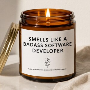 May include: A lit soy candle in an amber glass jar with a gold lid. The white label features the black text: SMELLS LIKE A BADASS SOFTWARE DEVELOPER, a small botanical line drawing, and the words MADE WITH ESSENTIAL OILS | HAND POURED SOY CANDLE.