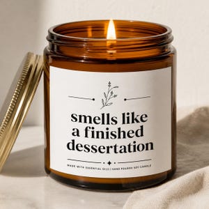 May include: A lit soy candle in an amber glass jar with a gold lid. The white label features a black floral graphic and the text: smells like a finished dissertation. Below the text, it reads: MADE WITH ESSENTIAL OILS | HAND POURED SOY CANDLE.