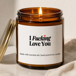 May include: A lit soy candle in a brown glass jar with a gold lid. The white label reads "I Fucking Love You" with a small red heart. The text "Made with essential oils | hand poured soy candle" is also on the label.