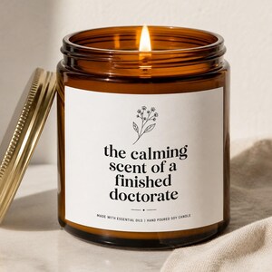 May include: A lit soy candle in a brown glass jar with a gold lid. The label reads "the calming scent of a finished doctorate." The candle is hand-poured and made with essential oils. A floral illustration is above the text.
