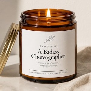 May include: A lit soy candle in a brown glass jar with a gold lid. The label reads "Smells Like A Badass Choreographer" with additional text about vision, grit, and creativity. The candle is hand-poured and made with essential oils.