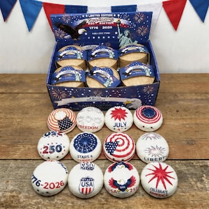 Puede incluir: Una exhibición de Liberty Dumplings con temática patriótica, con una caja con un águila y la Estatua de la Libertad. La colección incluye objetos redondos y decorativos con diseños de la bandera estadounidense, estrellas y textos como "Freedom" y "July 4th".