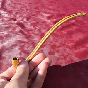 Op de afbeelding: Een lange, slanke, lichtgekleurde houten rookpijp met een gebogen steel en een kleine, ronde kop. De pijp heeft een opvallende donkerbruine lijn die over de lengte van de steel loopt en toont een natuurlijke houtnerfafwerking.