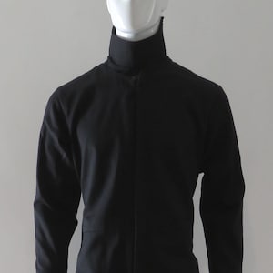 Può includere: Tuta nera con collo alto e maniche lunghe. L'indumento intero ha una cerniera a tutta lunghezza sul davanti ed è esposto su un manichino. La tuta è di colore uniforme e sembra essere realizzata in un tessuto resistente.