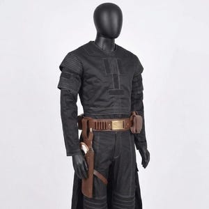 K&ouml;nnte beinhalten: Ein schwarzes Kost&uuml;m, das auf einer Schaufensterpuppe ausgestellt ist. Das Outfit umfasst ein lang&auml;rmliges Oberteil, eine Hose und einen G&uuml;rtel mit Taschen. Eine Halterung mit einer Waffe ist am Bein befestigt. Das Kost&uuml;m hat ein futuristisches Design mit sichtbaren N&auml;hten und gepolsterten Details.