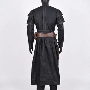 K&ouml;nnte beinhalten: Schwarzes Kost&uuml;m mit langem Mantel, lang&auml;rmliger Jacke und Hose. Die Jacke hat wei&szlig;e N&auml;hte und Schulterakzente. Ein brauner G&uuml;rtel und ein Holster werden um die Taille getragen. Das Outfit wird auf einer schwarzen Schaufensterpuppe pr&auml;sentiert.