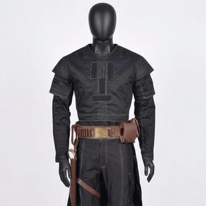 K&ouml;nnte beinhalten: Schwarzes Kost&uuml;m mit einem lang&auml;rmeligen Oberteil, einer Hose und einem Werkzeugg&uuml;rtel. Das Oberteil hat ein geometrisches Design mit wei&szlig;en N&auml;hten. Der G&uuml;rtel ist braun mit einer goldfarbenen Schnalle und einer braunen Tasche. Das Kost&uuml;m wird auf einer Schaufensterpuppe pr&auml;sentiert.