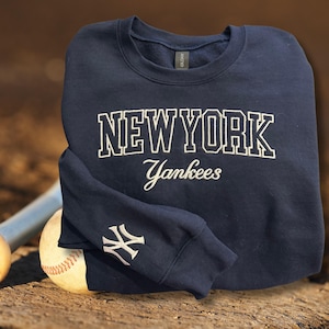 May include: Navy blue sweatshirt with the words "NEW YORK Yankees" in white block letters. The Yankees logo is on the left sleeve. A baseball and bat are in the background.
