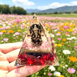 May include: A clear pyramid-shaped resin sculpture with a bronze-colored Ganesh statue inside. The pyramid has red and pink accents and sits in front of a field of colorful flowers.