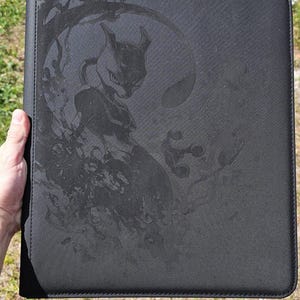 Puede incluir: Carpeta de cartas negra con una imagen en relieve detallada del personaje Mewtwo. La carpeta rectangular tiene un borde cosido y parece estar hecha de un material texturizado. El fondo está borroso, sugiriendo un entorno al aire libre.