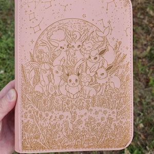 May include: A pink case with a gold illustration of cartoon creatures, a full moon, constellations, and floral elements. The case has a textured surface and a rectangular shape. The design is detailed and whimsical, suitable for fans of fantasy or animation.