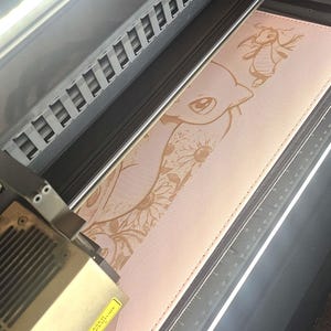 May include: A light pink rectangular mat featuring a laser-engraved design of a cartoon creature and floral patterns, positioned inside a laser engraving machine with a visible metal ruler along the side showing metric measurements.