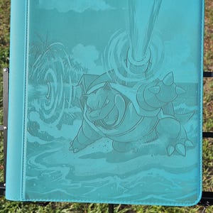 May include: A teal binder featuring an embossed illustration of a turtle-like creature in a water scene. The binder has a solid teal color with a smooth texture and a black metal stand.