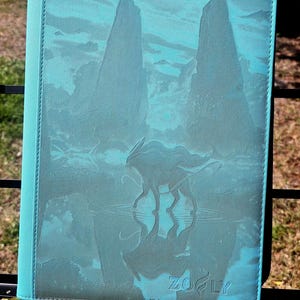 Puede incluir: Una funda de tableta turquesa con un diseño de paisaje que presenta una criatura y siluetas de montañas. La funda tiene un borde turquesa a juego y la marca "ZOFly" está impresa en la parte inferior.