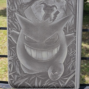 May include: A rectangular gray binder featuring a detailed laser-engraved illustration of the Pokémon Gengar in the foreground with a smaller Mimikyu figure positioned above it, set against a background of stylized trees and foliage.