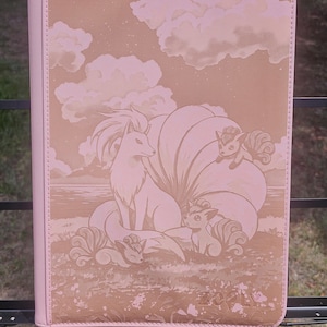 May include: Pink binder with a detailed illustration of a fox-like creature with multiple tails, set against a landscape with clouds. The design is in shades of pink and brown, with a stitched border.