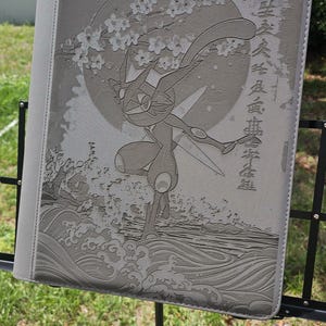 Puede incluir: Carpeta gris con una ilustración detallada de un personaje en una pose dinámica, sobre un fondo de luna llena, flores de cerezo y olas estilizadas. Texto japonés visible en el lado derecho de la carpeta.