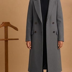Könnte beinhalten: Ein langer, grauer Doppelreiher-Mantel mit klassischem Reverskragen. Der Mantel hat zwei Knopfreihen und eine taillierte Passform. Die Person trägt ein schwarzes Oberteil und eine schwarze Hose. Ein hölzerner Kleiderständer und eine braune Ledertasche befinden sich im Hintergrund.