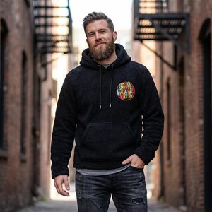 Puede incluir: Una sudadera con capucha negra con un gráfico circular colorido en el pecho. La sudadera tiene un bolsillo de canguro y una capucha con cordón. La persona lleva vaqueros grises y está de pie en un callejón.