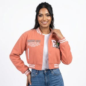 May include: Coral-colored cropped varsity jacket with white stripes on the collar, cuffs, and hem. The jacket features the text "RUNNIN RICH" and a graphic on the front. The jacket has a button-up closure.