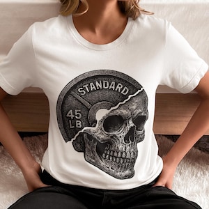 Puede incluir: Camiseta blanca con un diseño gráfico de una pesa y una calavera. La pesa lleva la inscripción "STANDARD" y "45 LB". La calavera está detallada con sombreado y líneas. La camiseta es de manga corta.