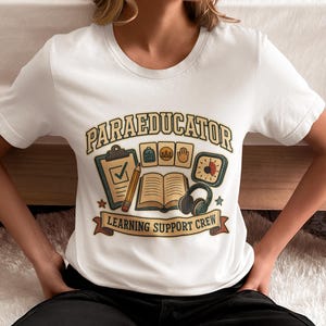 Puede incluir: Una camiseta blanca de manga corta con un gráfico que incluye el texto "PARAEDUCATOR" y "LEARNING SUPPORT CREW". El diseño muestra un portapapeles, un lápiz, un libro abierto, auriculares, un temporizador e iconos de mochila, personas y mano.