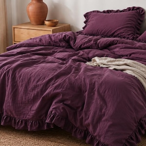 May include: A deep purple bedding set featuring a duvet cover and two matching pillowcases, all adorned with ruffled edges. A light beige throw blanket with a fringed hem is draped across the foot of the bed, adding texture to the ensemble.
