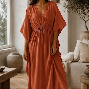 May include: A floor-length, rust-coloured kaftan dress featuring a deep V-neckline, wide kimono-style sleeves, and a drawstring waist with thin ties. The garment is made of a solid-coloured, lightweight fabric that falls in a loose, flowing silhouette.