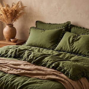 May include: An olive green bedding set featuring a duvet cover and matching pillowcases with ruffled edges. A light tan throw blanket is draped across the bottom of the bed. The bedding has a textured appearance and a solid dark green color.