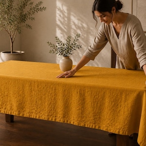 May include: A rectangular mustard yellow tablecloth with a visible woven texture draped over a wooden table. The fabric has a matte finish and hangs down the sides of the table, covering the surface completely with a consistent, solid color.