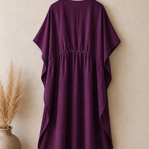 Puede incluir: Un vestido caft&aacute;n de color morado intenso con escote en V, mangas cortas anchas y una cintura el&aacute;stica fruncida. La prenda est&aacute; confeccionada en tela de color s&oacute;lido y cuelga de una percha de madera, mostrando una silueta holgada y fluida.