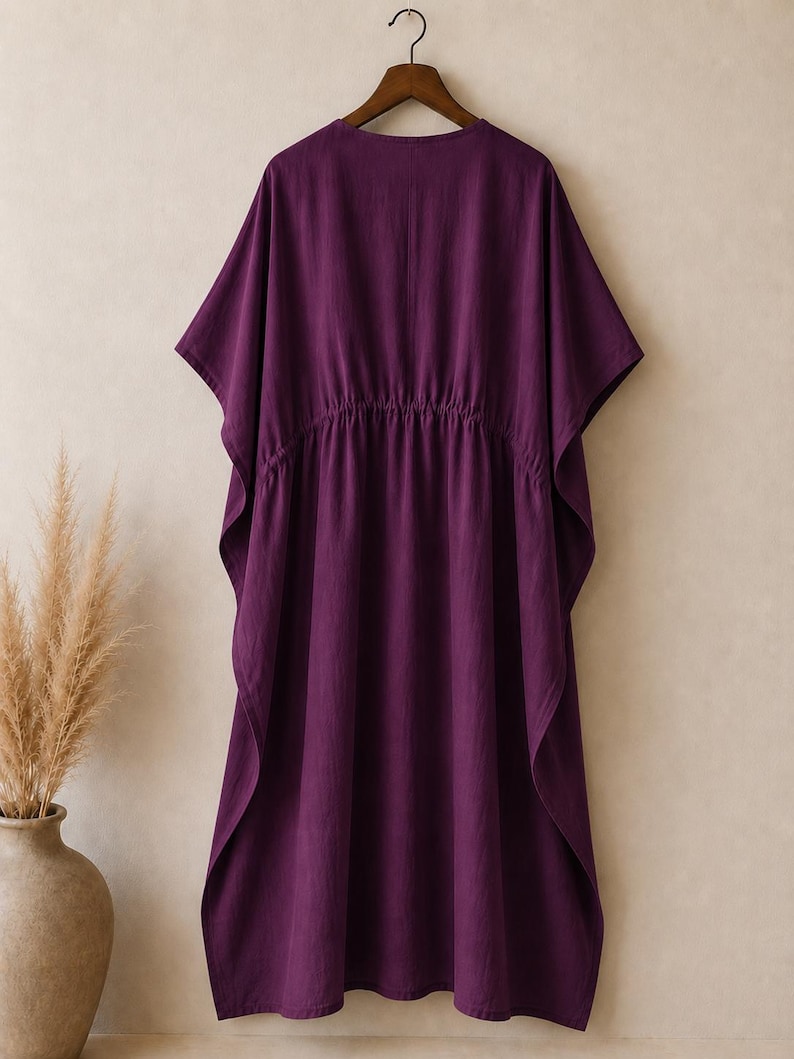 Puede incluir: Un vestido caft&aacute;n de color morado intenso con escote en V, mangas cortas anchas y una cintura el&aacute;stica fruncida. La prenda est&aacute; confeccionada en tela de color s&oacute;lido y cuelga de una percha de madera, mostrando una silueta holgada y fluida.