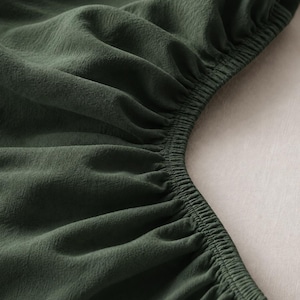 Op de afbeelding: Een donker bosgroen kledingstuk van stof met een geplooide, elastische halslijn. Het materiaal heeft een subtiele, fijnkorrelige textuur met zichtbare plooien en rimpels die door de elastische band worden gevormd, wat de diepe kleur benadrukt.