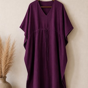 Puede incluir: Un vestido caft&aacute;n de color morado intenso con escote en V, mangas cortas anchas y cintura con cord&oacute;n ajustable y finas cintas a juego. La prenda se muestra en una percha de madera, exhibiendo una silueta holgada y fluida de tela mate.